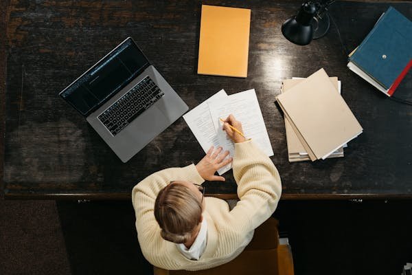 Overhead view of woman writing at desk with laptop, papers, and books.