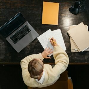 Overhead view of woman writing at desk with laptop, papers, and books.